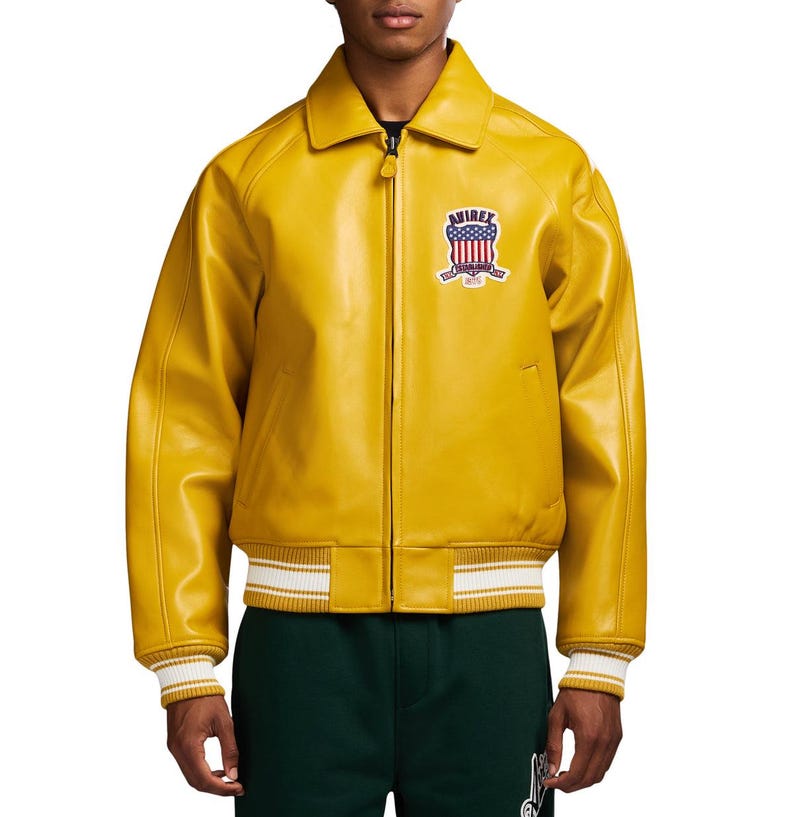May include: A mustard yellow leather bomber jacket with a pointed collar, a full-length front zipper, and ribbed cuffs and hem featuring white stripes. A patch on the left chest displays the text AVIREX above a shield with a red, white, and blue flag.