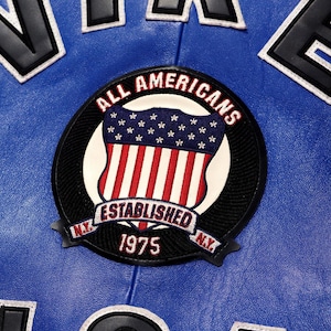 May include: A bright blue leather jacket featuring a circular embroidered patch. The patch displays a shield with stars and stripes, the text ALL AMERICANS, ESTABLISHED, 1975, and N.Y. The jacket also has large black and white lettering visible.