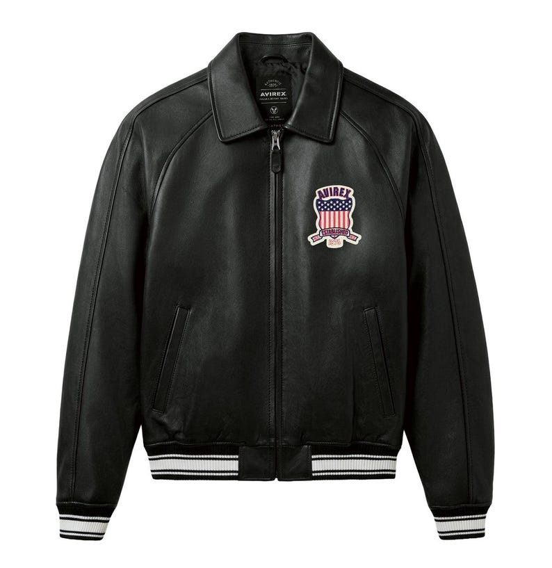 May include: A black leather jacket with a full-length front zip, a classic collar, and ribbed cuffs and hem featuring white stripes. A patch on the left chest displays the text AVIREX above a shield with a red, white, and blue flag design.
