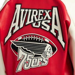 May include: A red and white varsity jacket featuring large white embroidered text that reads AVIREX USA. Below the text is a graphic of a football with wings and a profile of a person, accompanied by the white embroidered text 75ers.
