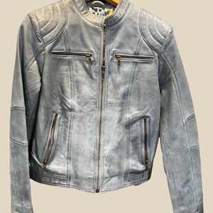 K&ouml;nnte beinhalten: Hellblaue Lederjacke mit Stehkragen und Rei&szlig;verschluss. Die Jacke hat mehrere Rei&szlig;verschlusstaschen und gesteppte Schulterdetails. Die Jacke h&auml;ngt an einem Holzkleiderb&uuml;gel.