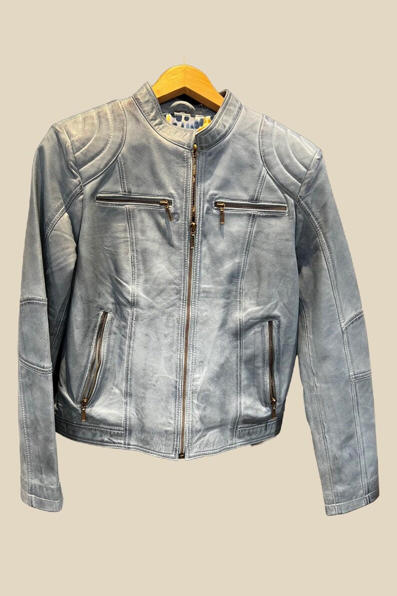 K&ouml;nnte beinhalten: Hellblaue Lederjacke mit Stehkragen und Rei&szlig;verschluss. Die Jacke hat mehrere Rei&szlig;verschlusstaschen und gesteppte Schulterdetails. Die Jacke h&auml;ngt an einem Holzkleiderb&uuml;gel.