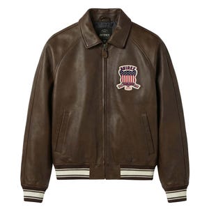 May include: A dark brown leather jacket with a front zip fastening, a classic collar, and ribbed cuffs and hem featuring white stripes. A patch on the left chest displays the text AVIREX above a shield graphic with a red, white, and blue flag design.