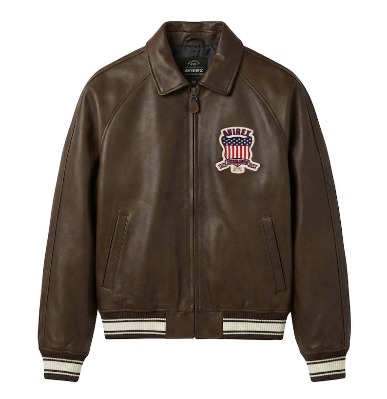 May include: A dark brown leather jacket with a front zip fastening, a classic collar, and ribbed cuffs and hem featuring white stripes. A patch on the left chest displays the text AVIREX above a shield graphic with a red, white, and blue flag design.
