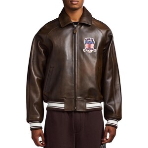May include: A dark brown leather bomber jacket featuring a front zip fastening, a pointed collar, and ribbed cuffs and hem with white stripes. A patch on the left chest displays the text AVIREX above a shield with a red, white, and blue flag design.