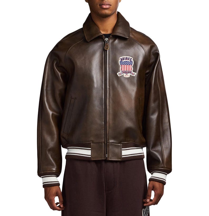 May include: A dark brown leather bomber jacket featuring a front zip fastening, a pointed collar, and ribbed cuffs and hem with white stripes. A patch on the left chest displays the text AVIREX above a shield with a red, white, and blue flag design.