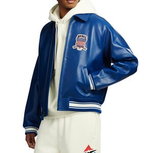 May include: A royal blue leather bomber jacket featuring a classic collar, long sleeves, and ribbed cuffs with white stripes. The chest displays a patch with the text AVIREX and a shield graphic. It is worn over a cream-coloured hooded sweatshirt.