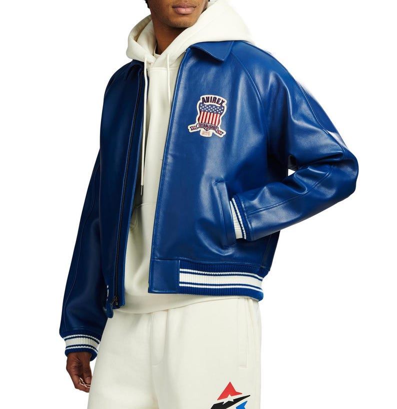 May include: A royal blue leather bomber jacket featuring a classic collar, long sleeves, and ribbed cuffs with white stripes. The chest displays a patch with the text AVIREX and a shield graphic. It is worn over a cream-coloured hooded sweatshirt.