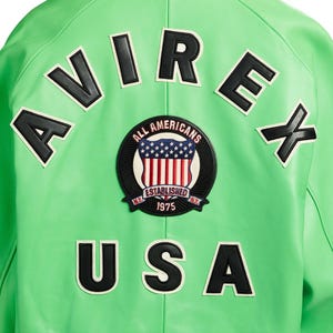 K&ouml;nnte beinhalten: Eine hell-limettengr&uuml;ne Jacke mit dem schwarzen Schriftzug AVIREX in einem Bogen oben und USA in schwarzen Buchstaben unten. Ein runder Aufn&auml;her in der Mitte zeigt einen Schild mit Sternen und Streifen, den Text ALL AMERICANS und 1975.