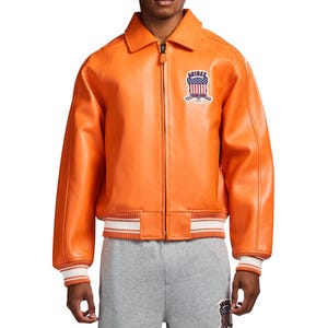 May include: A bright orange leather jacket with a full-length front zipper, a classic collar, and ribbed cuffs and hem featuring white stripes. On the left chest, there is a patch with the text AVIREX above a shield with stars and stripes.