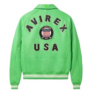 K&ouml;nnte beinhalten: Eine leuchtend hellgr&uuml;ne Lederjacke mit klassischem Kragen sowie gerippten B&uuml;ndchen und Saum mit wei&szlig;en Streifen. Auf der R&uuml;ckseite befinden sich der schwarze Schriftzug AVIREX und USA sowie ein rundes Abzeichen mit US-Flagge und dem Text ALL AMERICANS ESTABLISHED 1975.