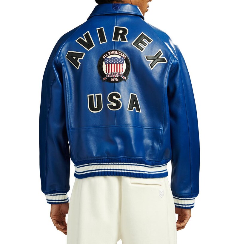 May include: A vibrant blue leather varsity jacket featuring the text AVIREX in large black letters across the upper back. Below the text is a circular patch with the American flag and the words ALL AMERICANS ESTABLISHED 1975. USA is printed in black below the patch.