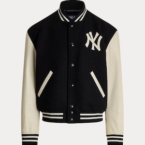 Op de afbeelding: Zwarte en crèmekleurige varsity-jas met het New York Yankees-logo. De jas heeft crèmekleurige mouwen, een zwarte body en gestreepte details op de manchetten en zoom. Het heeft een knoopsluiting en een geribbelde kraag.