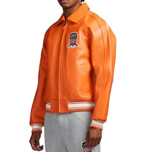 May include: A bright orange leather jacket featuring a classic collar, a front zipper closure, and ribbed cuffs and hem with white stripes. A patch on the chest displays the text AVIREX with a graphic of the American flag and eagle emblem.