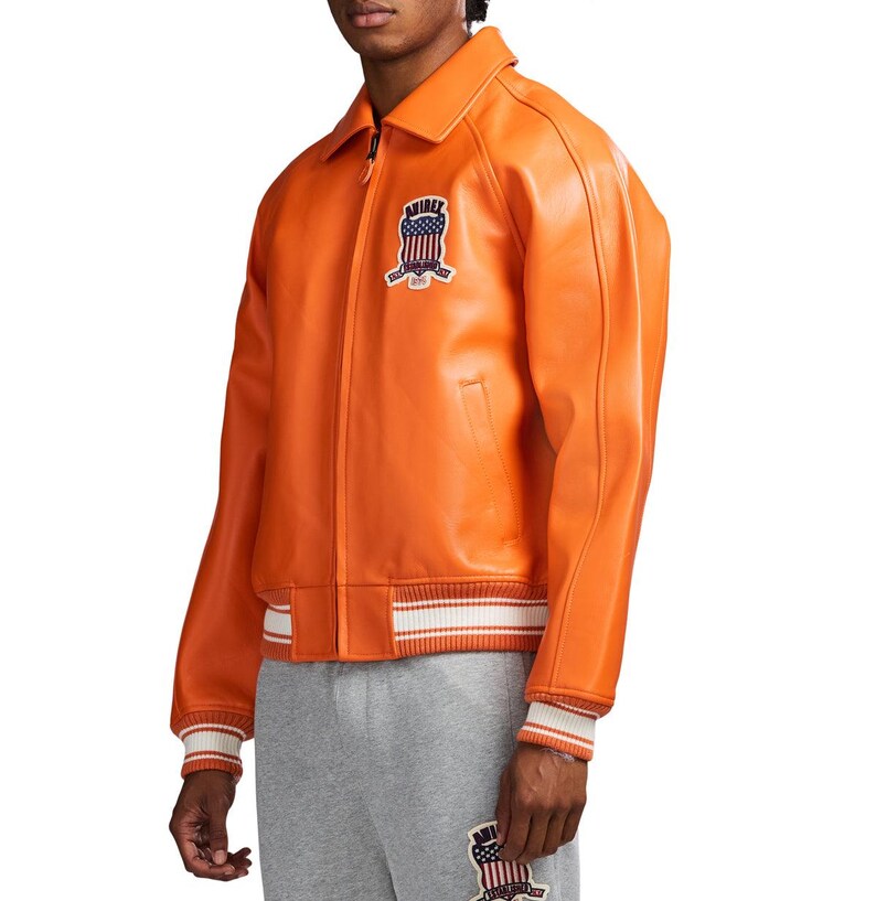 May include: A bright orange leather jacket featuring a classic collar, a front zipper closure, and ribbed cuffs and hem with white stripes. A patch on the chest displays the text AVIREX with a graphic of the American flag and eagle emblem.