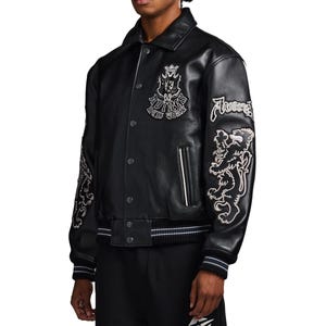 May include: A black leather varsity jacket featuring a pointed collar, snap button closure, and ribbed cuffs with white stripes. The chest displays an embroidered crest with the text "AVIREX NEW YORK," while the sleeves feature large lion patches.