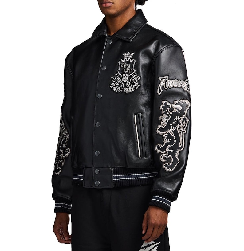 May include: A black leather varsity jacket featuring a pointed collar, snap button closure, and ribbed cuffs with white stripes. The chest displays an embroidered crest with the text "AVIREX NEW YORK," while the sleeves feature large lion patches.
