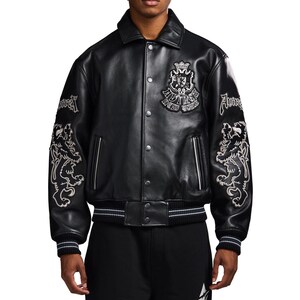 May include: A black leather varsity jacket featuring a pointed collar, snap-button front closure, and ribbed cuffs and hem with white stripes. The chest displays a crest patch with the text "AVIREX NEW YORK," while the sleeves feature lion embroidery.