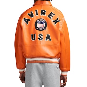 May include: An orange leather varsity jacket featuring a collar and ribbed cuffs with white stripes. The back displays the black text AVIREX and USA, along with a circular patch showing an American flag and the text ALL AMERICANS ESTABLISHED 1975.