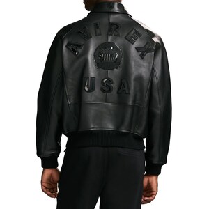 Puede incluir: Una chaqueta bomber de cuero negro con cuello, pu&ntilde;os y dobladillo acanalados. La parte posterior muestra el texto AVIREX en formaci&oacute;n arqueada, un emblema circular con un escudo estrellado y las letras USA en una fuente negra en relieve.