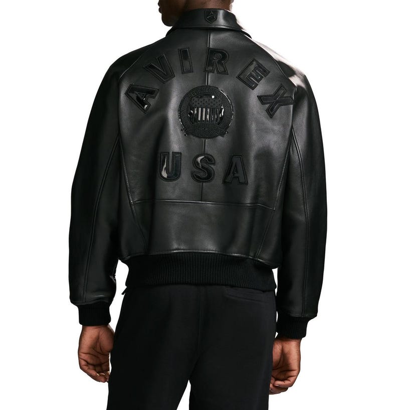 Puede incluir: Una chaqueta bomber de cuero negro con cuello, pu&ntilde;os y dobladillo acanalados. La parte posterior muestra el texto AVIREX en formaci&oacute;n arqueada, un emblema circular con un escudo estrellado y las letras USA en una fuente negra en relieve.