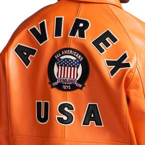 May include: A bright orange leather jacket featuring the black text AVIREX arched across the upper back. A circular patch in the center displays the text ALL AMERICANS, ESTABLISHED 1975, and N.Y. with a shield graphic. The black text USA is below.
