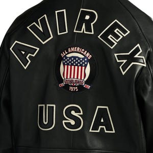 May include: A black leather jacket featuring the text AVIREX in large white and black letters across the top back. A circular patch in the centre displays the American flag with the text ALL AMERICANS ESTABLISHED 1975 N.Y. The word USA is below.