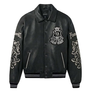 May include: A black leather jacket featuring a classic collar, snap button closure, and two front pockets. The chest displays a crest patch with the text Avirex New York. Each sleeve features an embroidered lion graphic and the word Avirex.