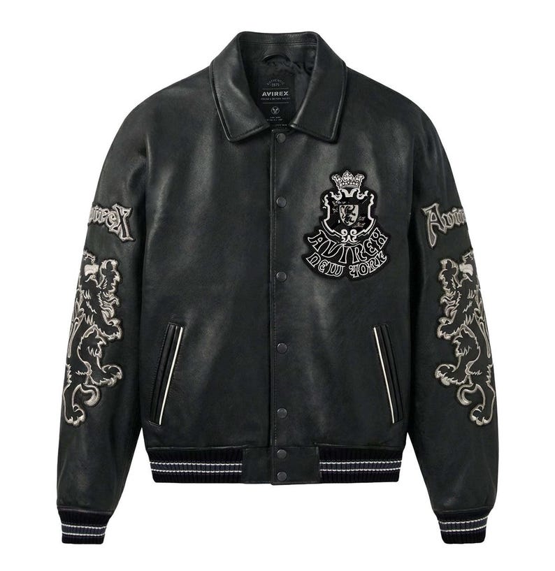 May include: A black leather jacket featuring a classic collar, snap button closure, and two front pockets. The chest displays a crest patch with the text Avirex New York. Each sleeve features an embroidered lion graphic and the word Avirex.