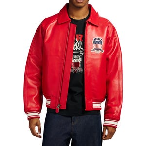 May include: A bright red leather jacket featuring a classic collar, a front zipper closure, and ribbed cuffs with white stripes. An embroidered patch with the text Avirex and a shield graphic is visible on the left chest area of the jacket.