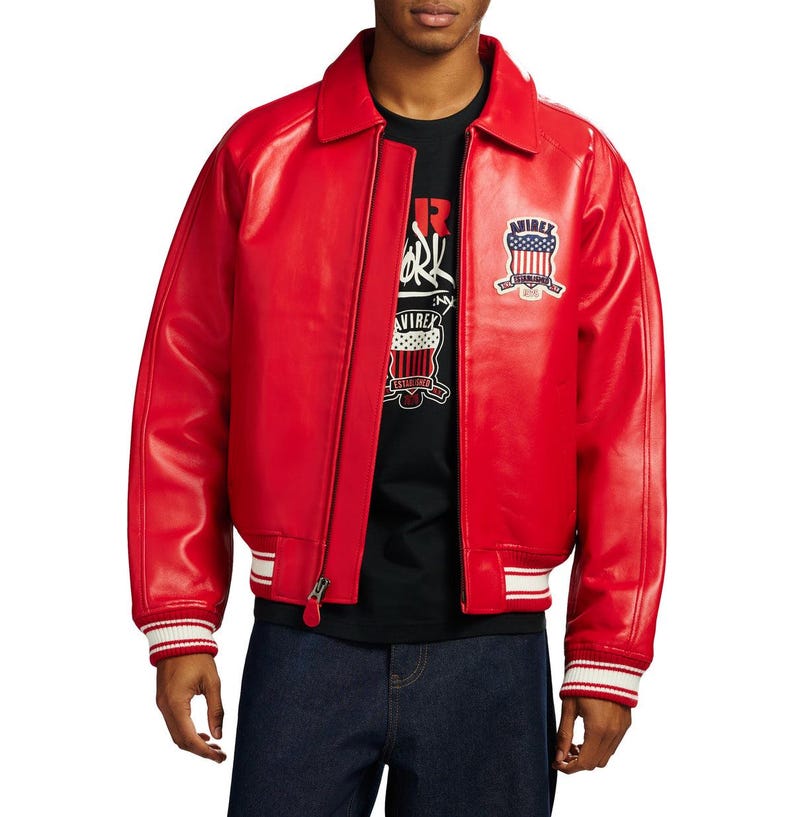 May include: A bright red leather jacket featuring a classic collar, a front zipper closure, and ribbed cuffs with white stripes. An embroidered patch with the text Avirex and a shield graphic is visible on the left chest area of the jacket.