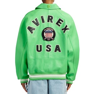 K&ouml;nnte beinhalten: Eine leuchtend hellgr&uuml;ne Leder-Collegejacke mit dem Schriftzug AVIREX in schwarzen Buchstaben, der &uuml;ber einem runden Aufn&auml;her mit US-Flagge und dem Text ALL AMERICANS ESTABLISHED 1975 gew&ouml;lbt ist, sowie dem schwarzen Schriftzug USA darunter.
