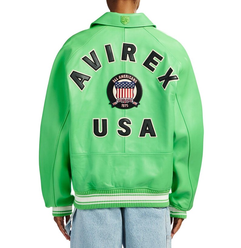K&ouml;nnte beinhalten: Eine leuchtend hellgr&uuml;ne Leder-Collegejacke mit dem Schriftzug AVIREX in schwarzen Buchstaben, der &uuml;ber einem runden Aufn&auml;her mit US-Flagge und dem Text ALL AMERICANS ESTABLISHED 1975 gew&ouml;lbt ist, sowie dem schwarzen Schriftzug USA darunter.