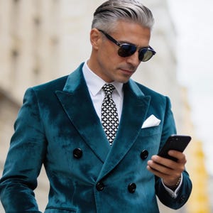 May include: A teal velvet double-breasted blazer with black buttons, worn over a white shirt and a black and white patterned tie. The person is wearing sunglasses and holding a black smartphone.