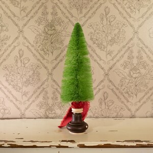 May include: A small, green, conical Christmas tree with a bottle brush texture. The tree sits atop a dark brown base with a red fabric accent. The background features a floral patterned wallpaper.