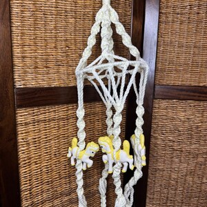 May include: A cream-colored macrame hanging mobile featuring twisted rope strands and a central diamond-shaped structure. Three small, white and yellow ceramic carousel horse figures are attached to the lower sections of the hanging ropes.
