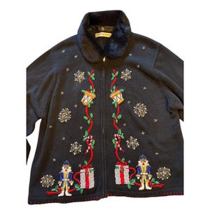 May include: Black zip-up sweater with a faux fur collar. The sweater features embroidered Christmas-themed designs, including snowflakes, bells, gift boxes, and nutcracker figures. The design incorporates red, green, gold, and white accents.
