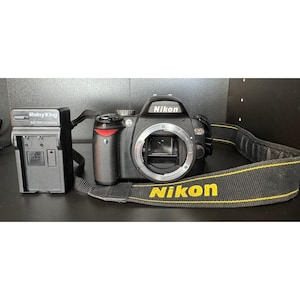 May include: A black Nikon D90 digital SLR camera with a yellow and black Nikon branded strap. A black battery charger is to the left of the camera. The camera lens is open.
