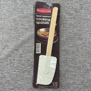 Peut inclure: Spatule de cuisine résistante à la chaleur Rubbermaid. Elle possède une tête rectangulaire blanche et un manche en bois clair. L'emballage indique "spatule de cuisine résistante à la chaleur" et la mesure "in long (26,6 cm)."
