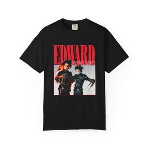 Könnte beinhalten: Ein schwarzes Kurzarm-T-Shirt mit einem Grafikdruck des Films Edward Scissorhands. Das Design zeigt den roten Schriftzug EDWARD SCISSORHANDS über einem fotografischen Bild von zwei Charakteren in schwarzen Outfits mit scherenartigen Händen.