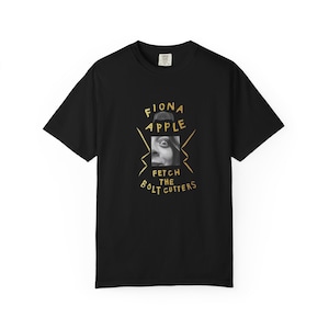 Op de afbeelding: Een zwart t-shirt met korte mouwen en een grafische print met de tekst FIONA APPLE en FETCH THE BOLT CUTTERS in het geel. Het ontwerp bevat een vierkante zwart-witfoto van een gezicht, gecentreerd tussen de regels gele tekst.