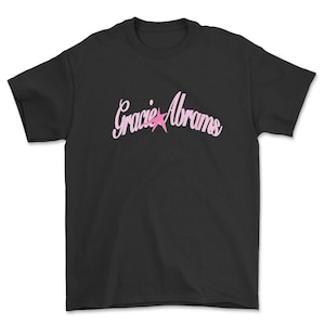 Op de afbeelding: Zwart t-shirt met de naam "Gracie Abrams" in lichtroze cursief schrift en een roze ster. Het t-shirt heeft een ronde hals.