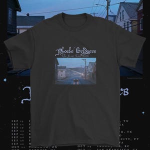 Puede incluir: Una camiseta gris carbón de manga corta con el texto Phoebe Bridgers The Tour 2026 en una fuente estilizada sobre una impresión fotográfica rectangular de una escena callejera al atardecer con un coche y casas bajo un cielo azul.