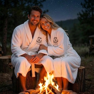 May include: A couple wearing white terry cloth robes with embroidered logos, sitting on a tree stump near a campfire. The robes have a shawl collar and a matching belt. The background includes a starry night sky and trees.