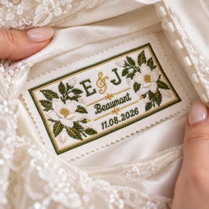 May include: A rectangular white fabric patch with pinked edges, featuring embroidered white magnolia flowers with green leaves. The text reads E & J, Beaumont, and 11.08.2026 in dark green and gold thread, centered on a satin garment.