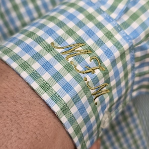 May include: Close-up of a shirt cuff with a blue, green, and white checkered pattern. The cuff features embroidered initials in a gold color. The shirt is made of a lightweight fabric.