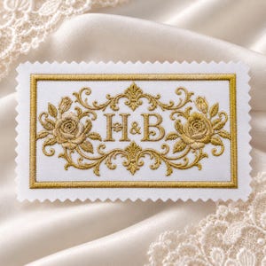 May include: A rectangular white fabric patch with pinked edges featuring gold embroidery. The design includes the letters H&B centered between two detailed gold embroidered roses, all enclosed within a decorative gold border frame.