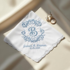 May include: White handkerchief with a scalloped edge, embroidered with a blue floral design and the letter "B". The names "James & Lauren" and the date "12.01.2025" are embroidered below the design. Gold earrings and a ring are also present.