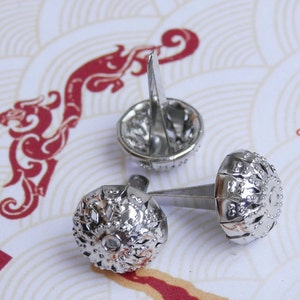 May include: Three silver metal decorative tacks with a detailed design. The tacks have a round head with a pointed end.