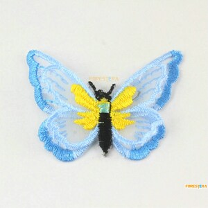 May include: A blue and yellow embroidered butterfly applique with a white mesh background. The butterfly has a black body and yellow wings with a blue outline.